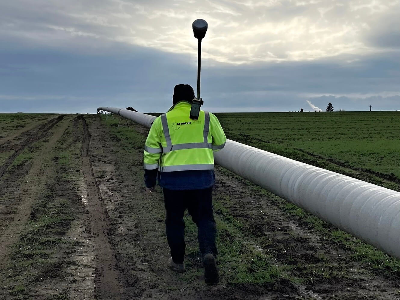 Technik v reflexní vestě kontroluje potrubí v zemědělské krajině pod zamračenou oblohou.